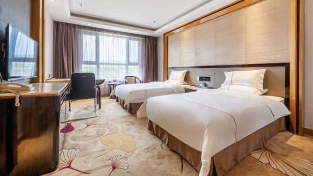 Ein Hotelzimmer mit zwei Betten, einem Schreibtisch, einem Stuhl, einem Fernseher und einem großen Fenster mit Vorhängen.