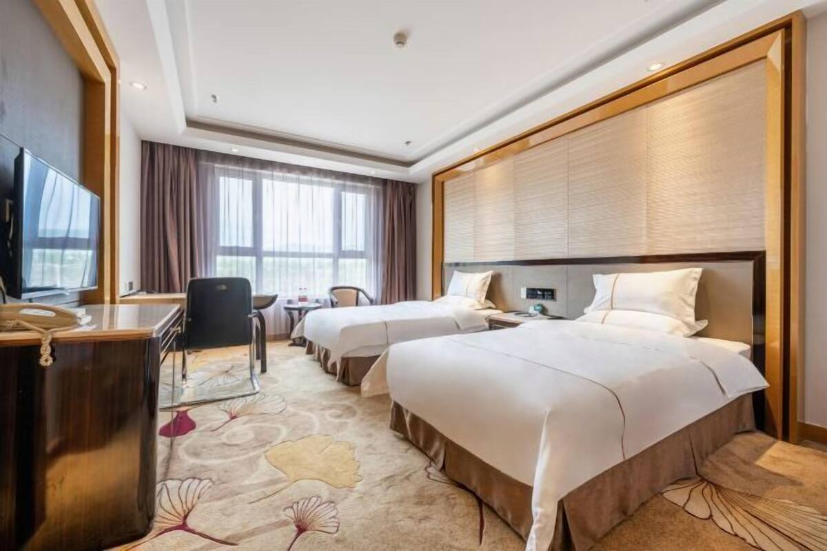 Ein Hotelzimmer mit zwei Betten, einem Schreibtisch, einem Stuhl, einem Fernseher und einem großen Fenster mit Vorhängen.