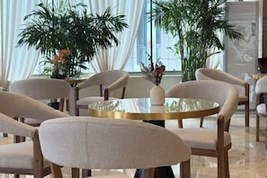 Uma área de refeições moderna com mesas e cadeiras redondas, plantas em vasos grandes e vista para a cidade através de janelas amplas.