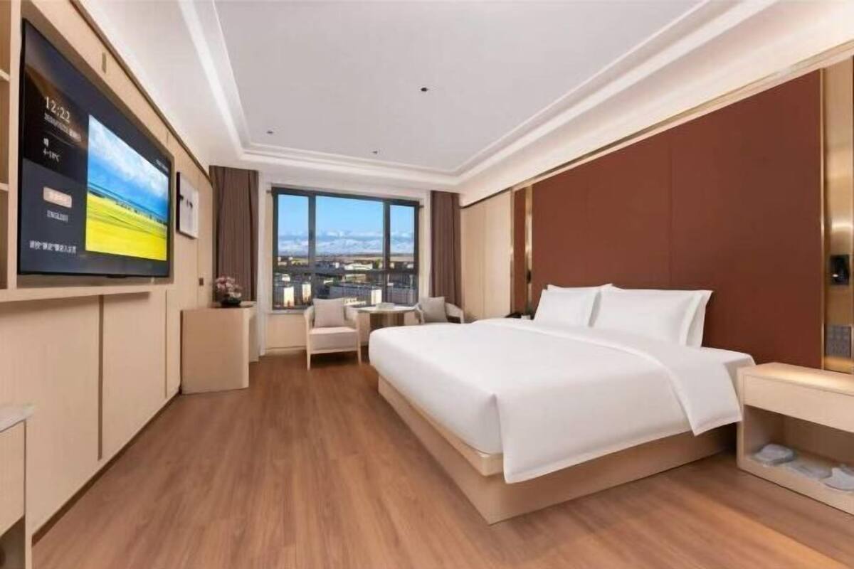 Ein Hotelzimmer mit einem großen Bett, einem an der Wand montierten Fernseher, einem kleinen Tisch und Blick auf die Stadt durch große Fenster.