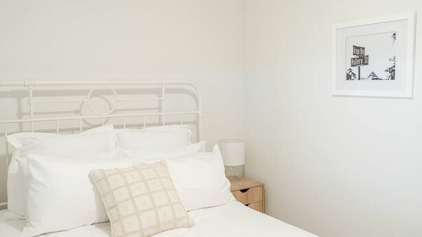 Un lit blanc avec une tête de lit en métal, une literie blanche et une petite table de chevet avec une lampe.