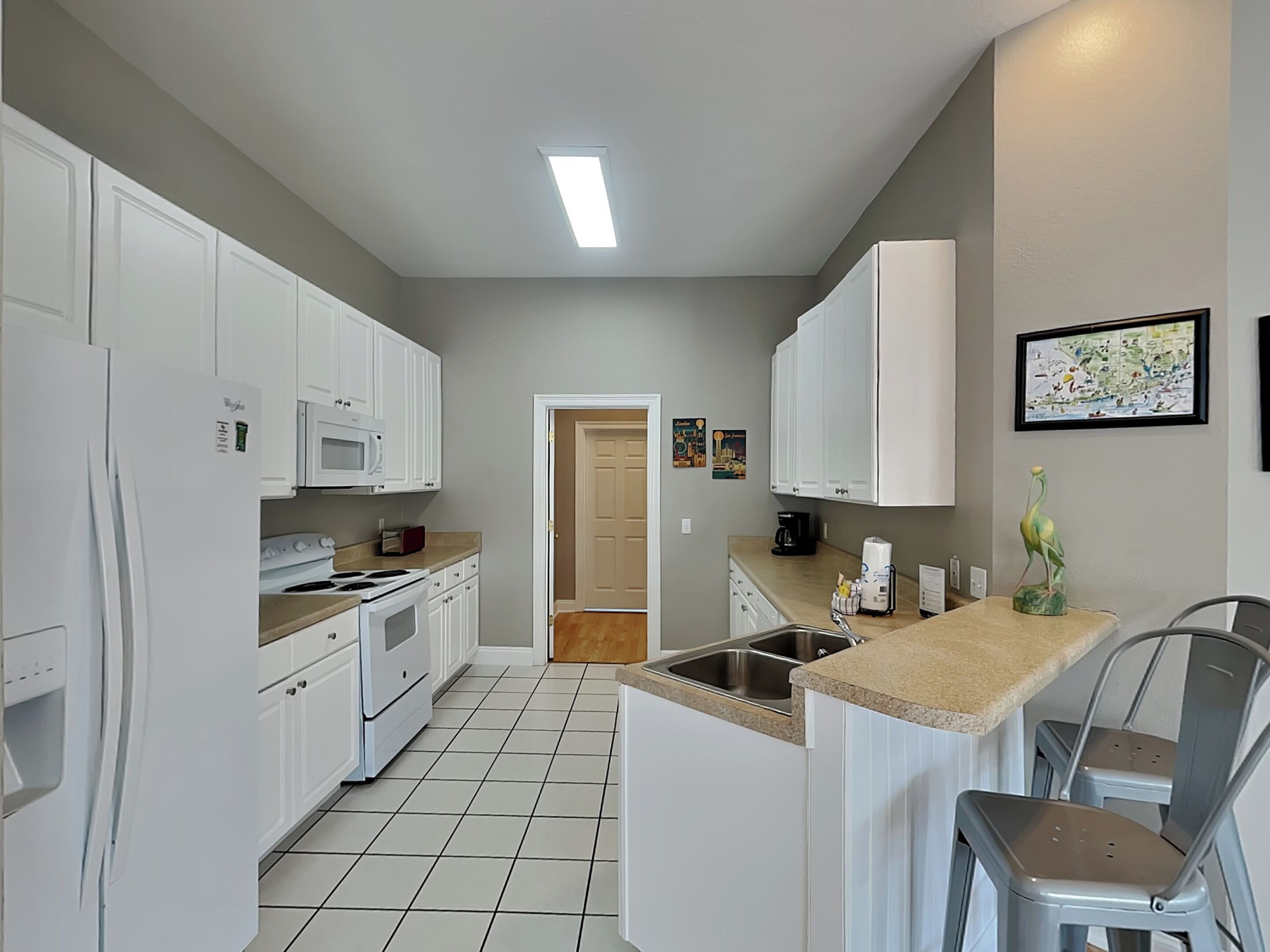 Een compacte keuken met witte kastjes, een magnetron, een fornuis en een spoelbak. Een deur leidt naar een andere kamer, en een klein eiland met een spoelbak en een stoel.