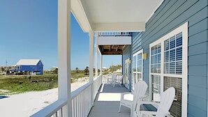 En overdækket veranda med hvide stole, udsigt til stranden og et blåt hus i baggrunden.