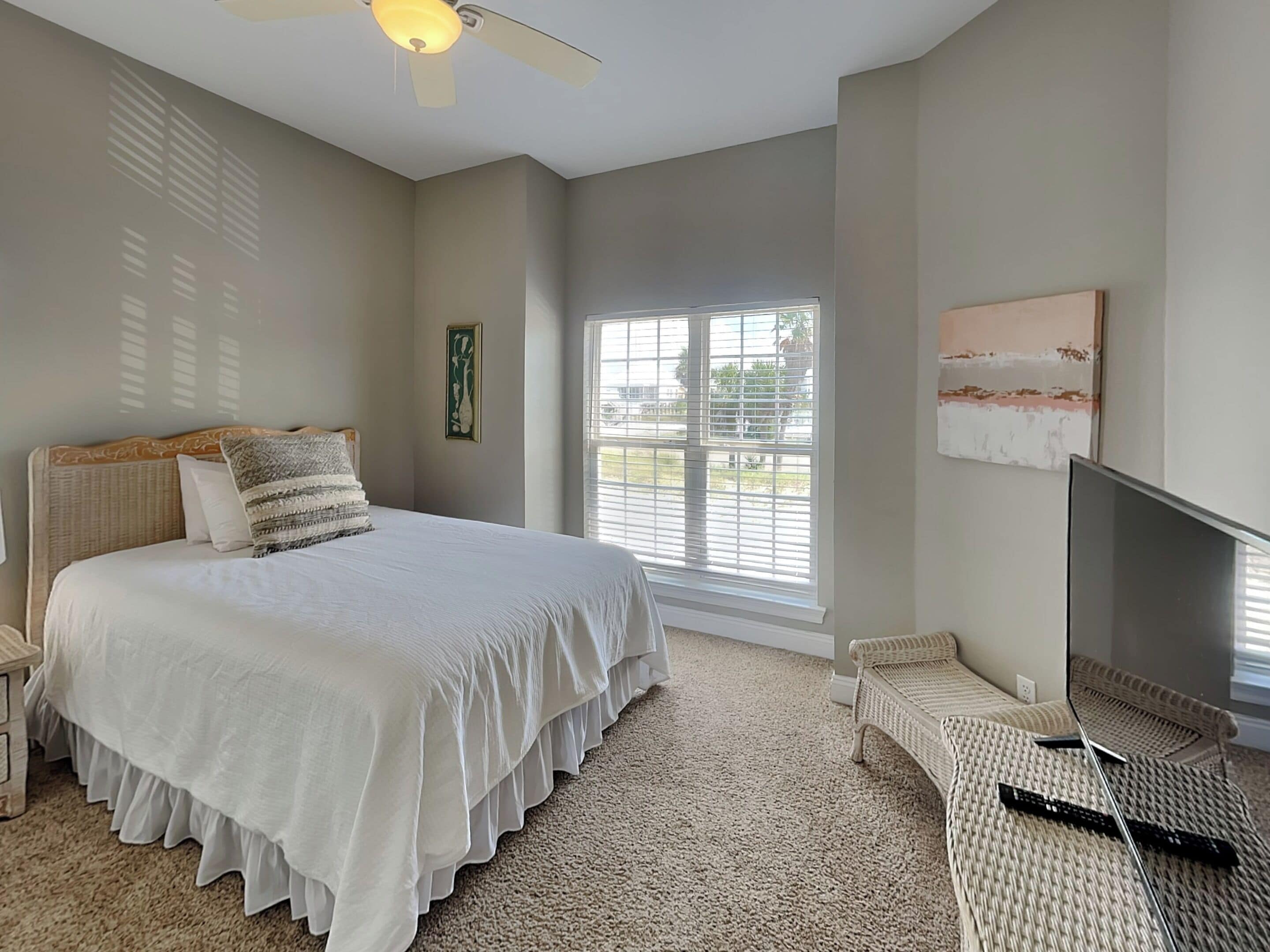 A bedroom with a large bed, a ceiling fan, a television, and a window with blinds.