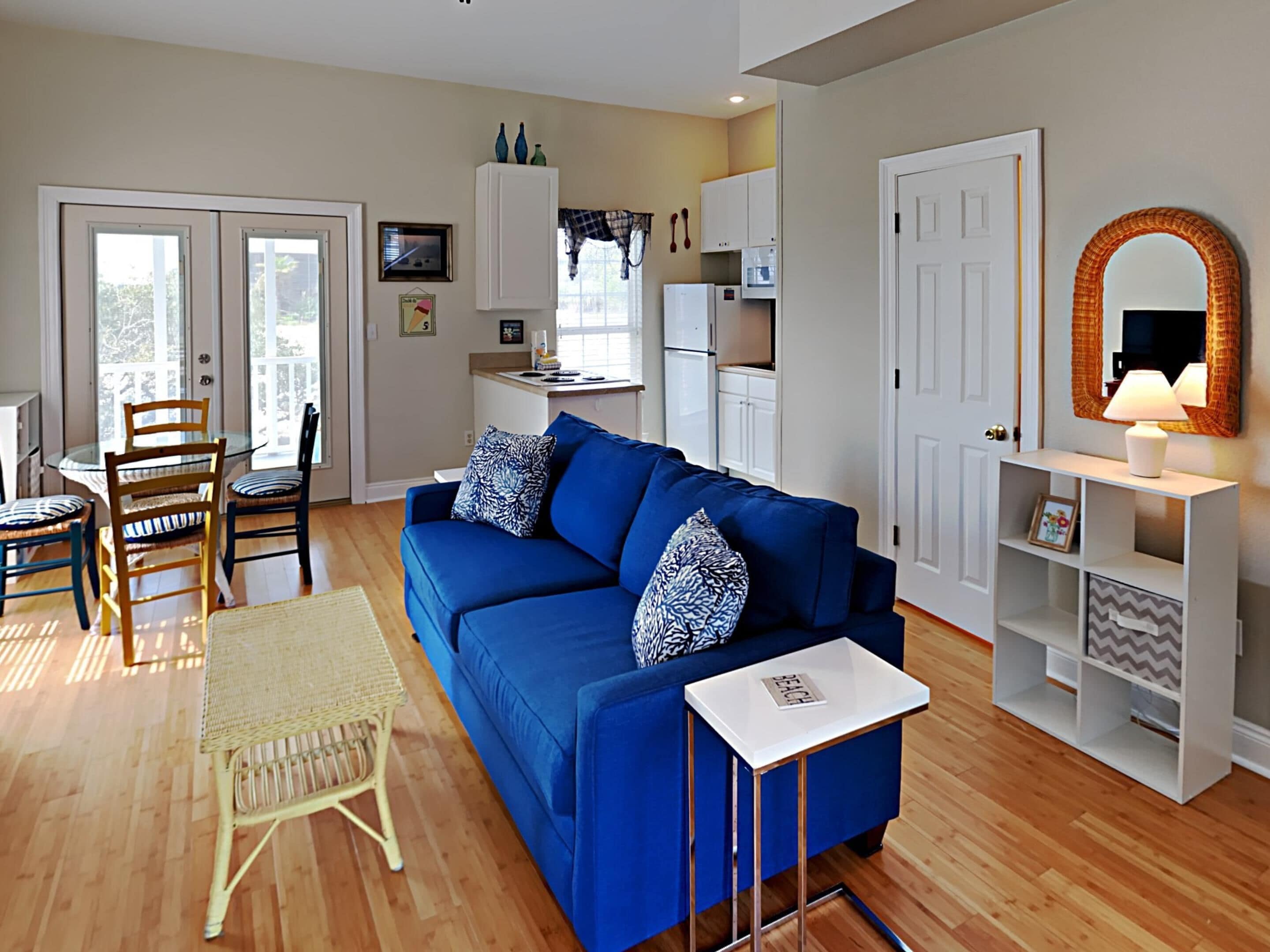 A living room with a blue sofa, a small white table, and a wicker chair. There is a kitchen area with cabinets and a refrigerator in the background.