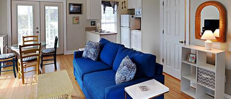 A living room with a blue sofa, a small white table, and a wicker chair. There is a kitchen area with cabinets and a refrigerator in the background.