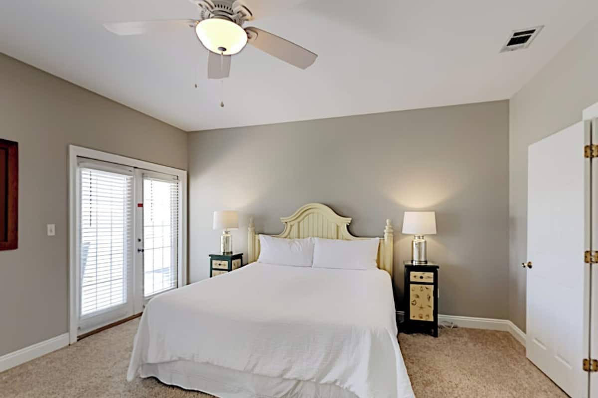 A bedroom with a bed, two bedside tables with lamps, a ceiling fan, a door, and a window with blinds.