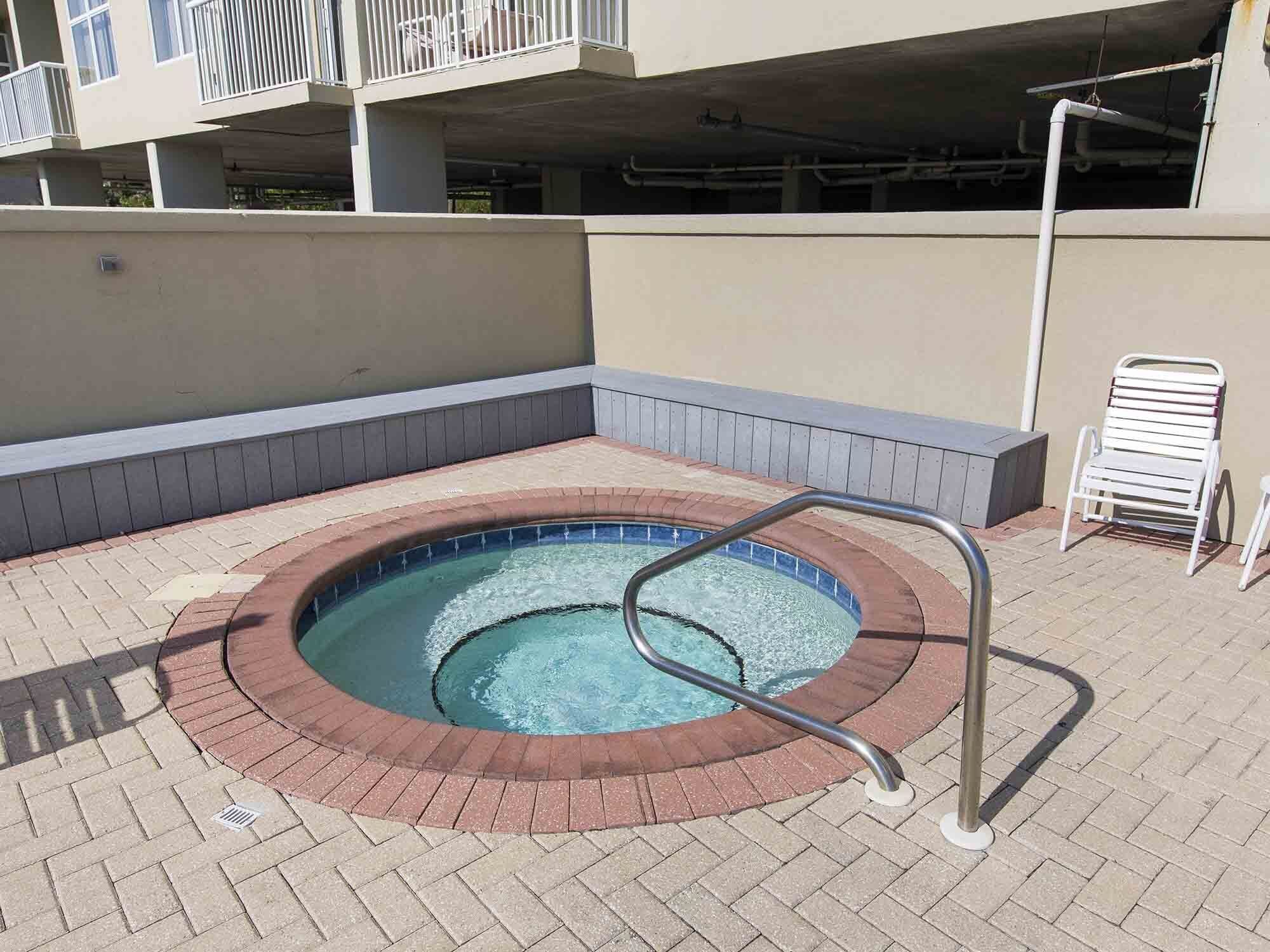 Un jacuzzi con escalera metálica, rodeado por un borde de ladrillo, en un espacio exterior con un edificio al fondo.
