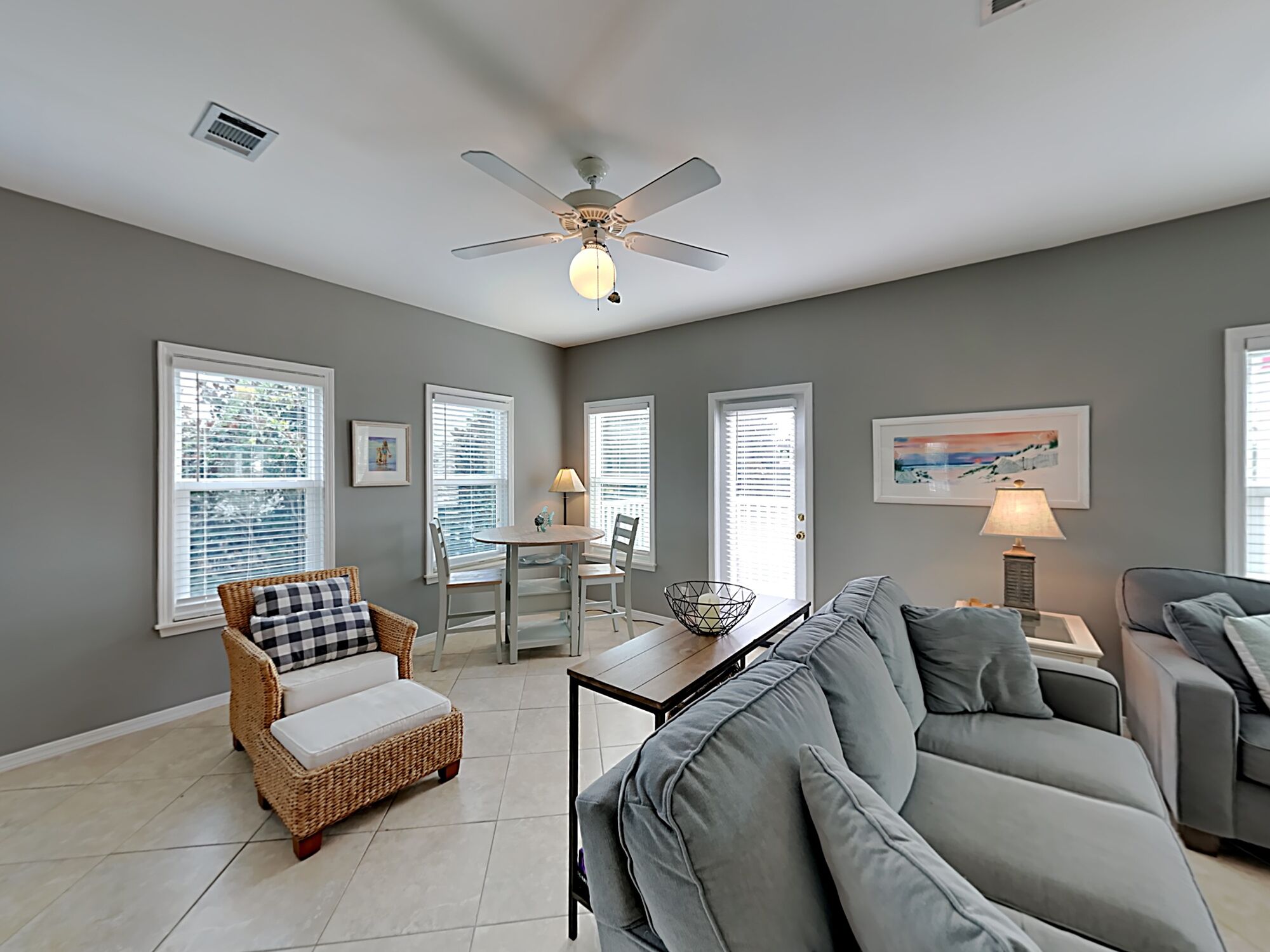 A spacious living room with a grey couch, wicker chair, dining table, and ceiling fan.