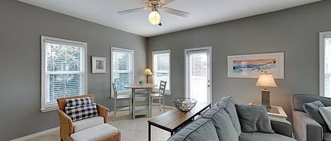 A spacious living room with a grey couch, wicker chair, dining table, and ceiling fan.