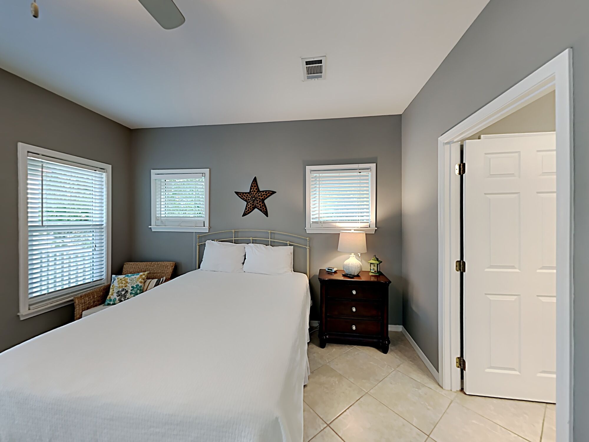 A bedroom with a bed, two windows with blinds, a star decoration, a nightstand with a lamp, and a door.