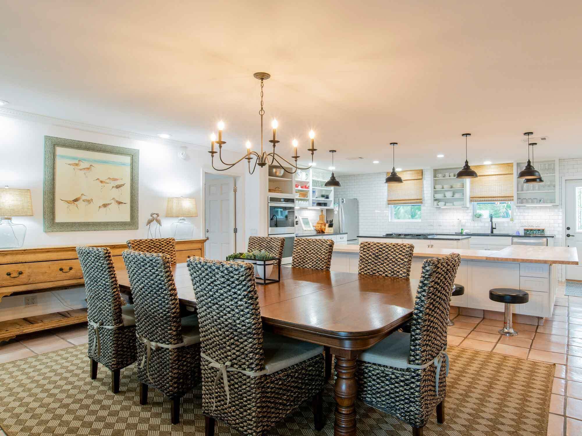 A dining area with a wooden table and woven chairs, a chandelier, and a kitchen in the background.