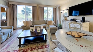 A spacious living room with a sofa, coffee table, and a dining table. There is a ceiling fan, large windows with curtains, and a flat-screen TV mounted on the wall. - Gulf Place Six Palms #2B - BB's Hideaway by Southern Vacation Rentals (Santa Rosa Beach)
