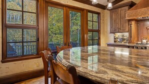 A kitchen with a large island, wooden cabinets, and a view of trees through large windows. - 5 Beds in BC - Ski In, Hot Tub, Pool (Beaver Creek)