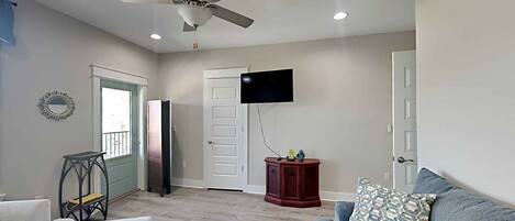 A living room with a ceiling fan, a flat-screen TV mounted on the wall, a grey sofa with patterned cushions, a small wooden side table, and a white chair.
