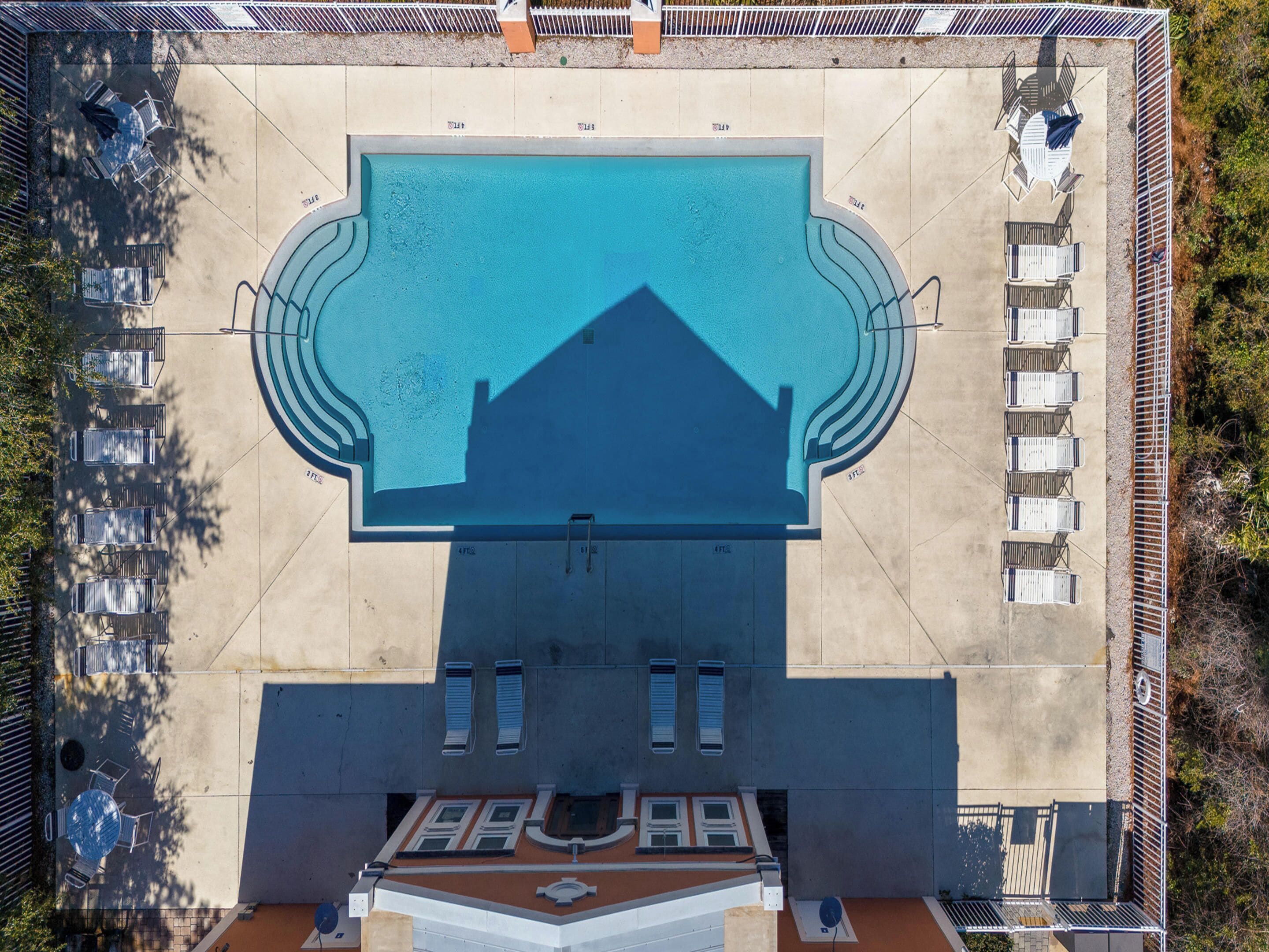 Eine Vogelperspektive zeigt einen Swimmingpool mit geometrischem Design, umgeben von einem Zaun und mehreren Sitzbereichen.