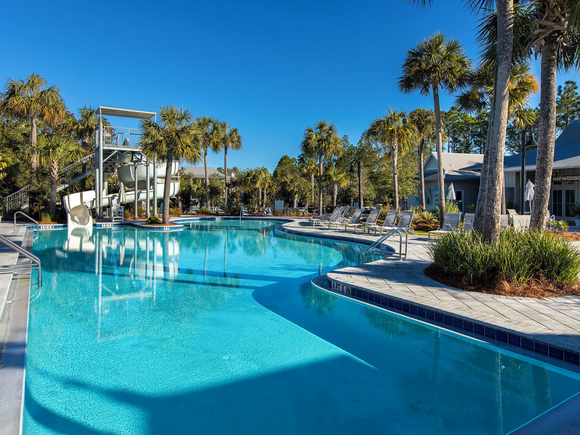 A clear blue pool with a water slide, surrounded by palm trees and lounge chairs.