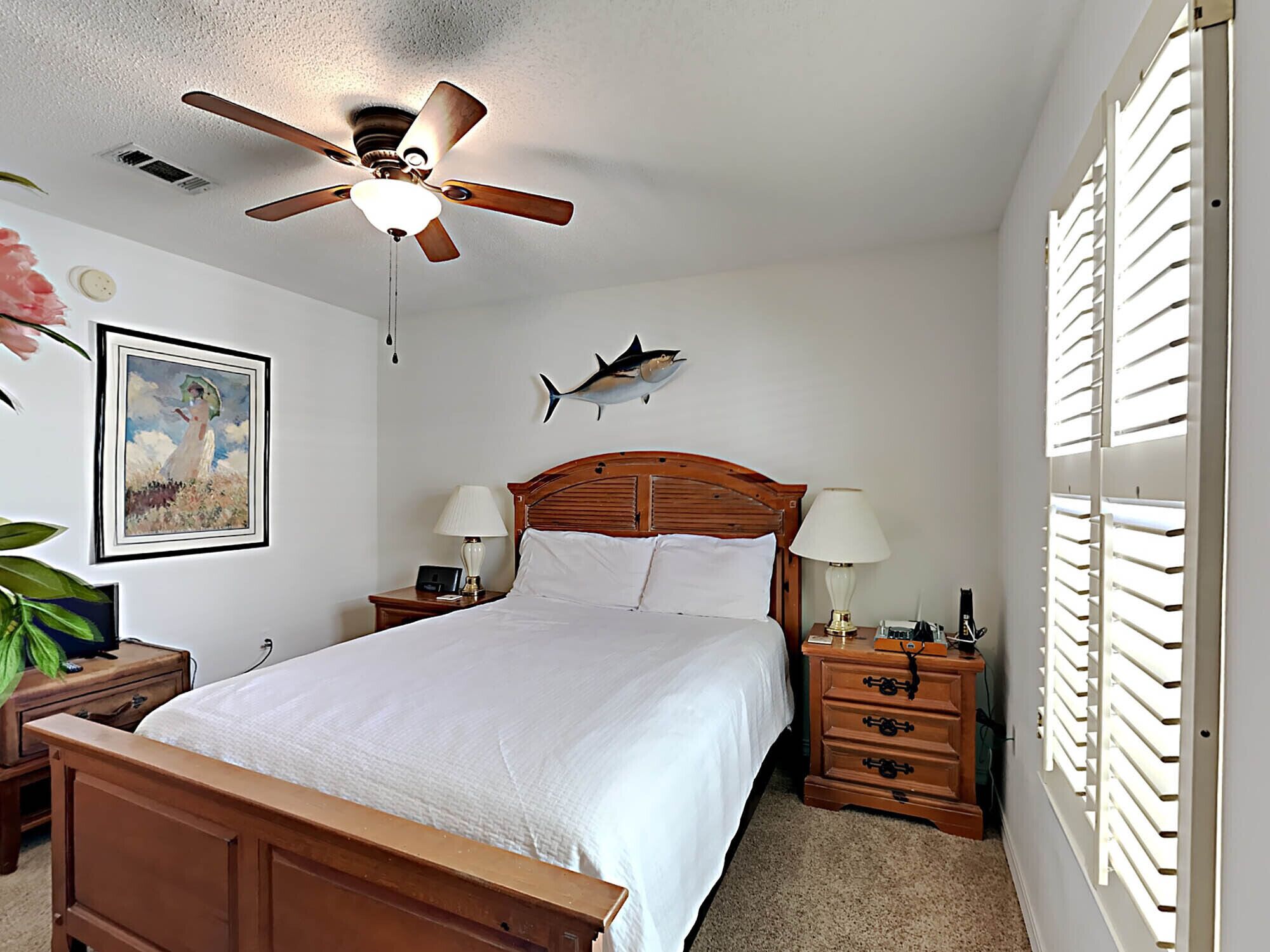 A bedroom with a wooden bed, two bedside tables with lamps, a ceiling fan, a framed painting, and a wall-mounted shark decoration.