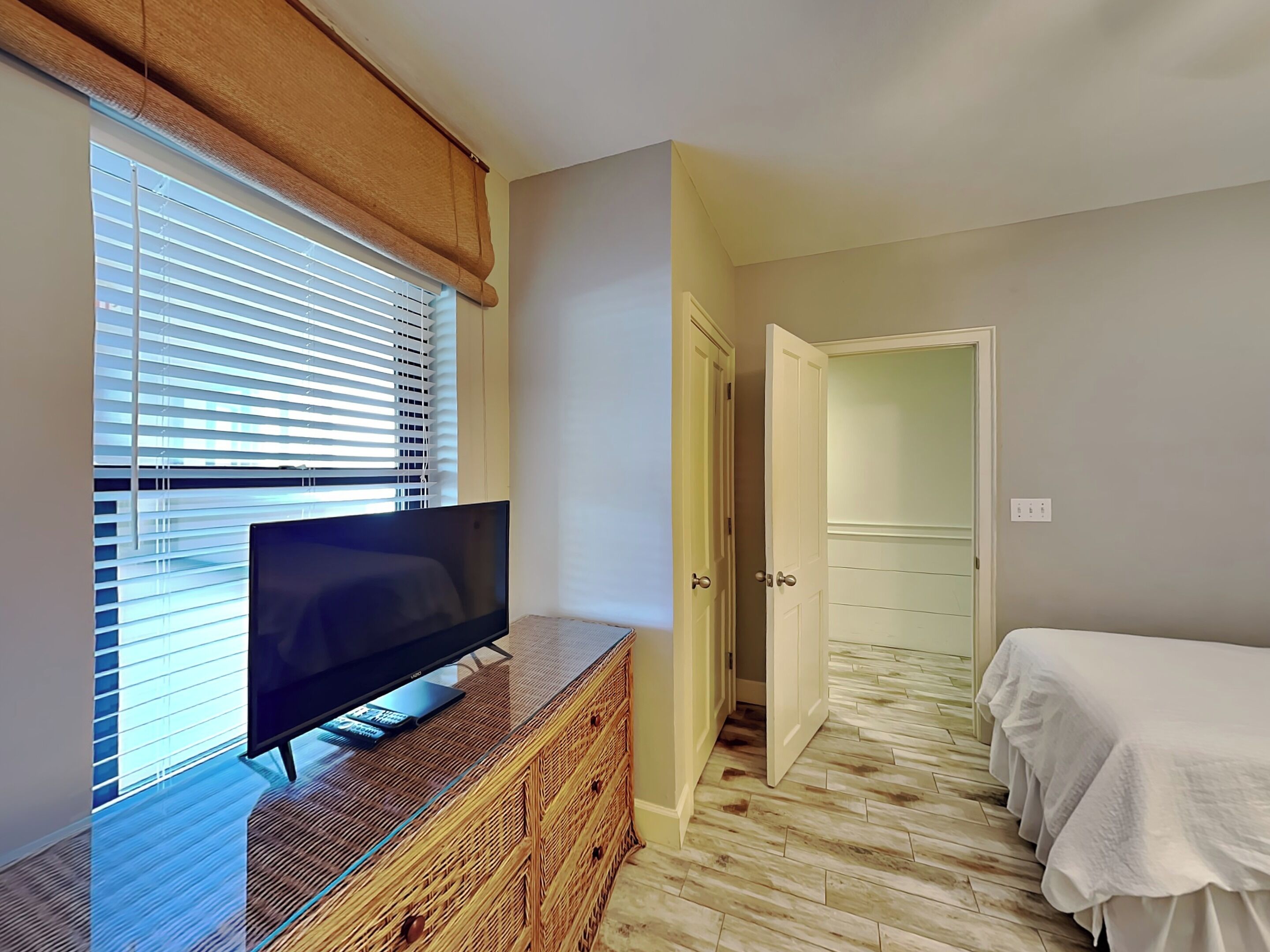 A bedroom with a TV on a wooden dresser, a bed with a white cover, and a door leading to another room.