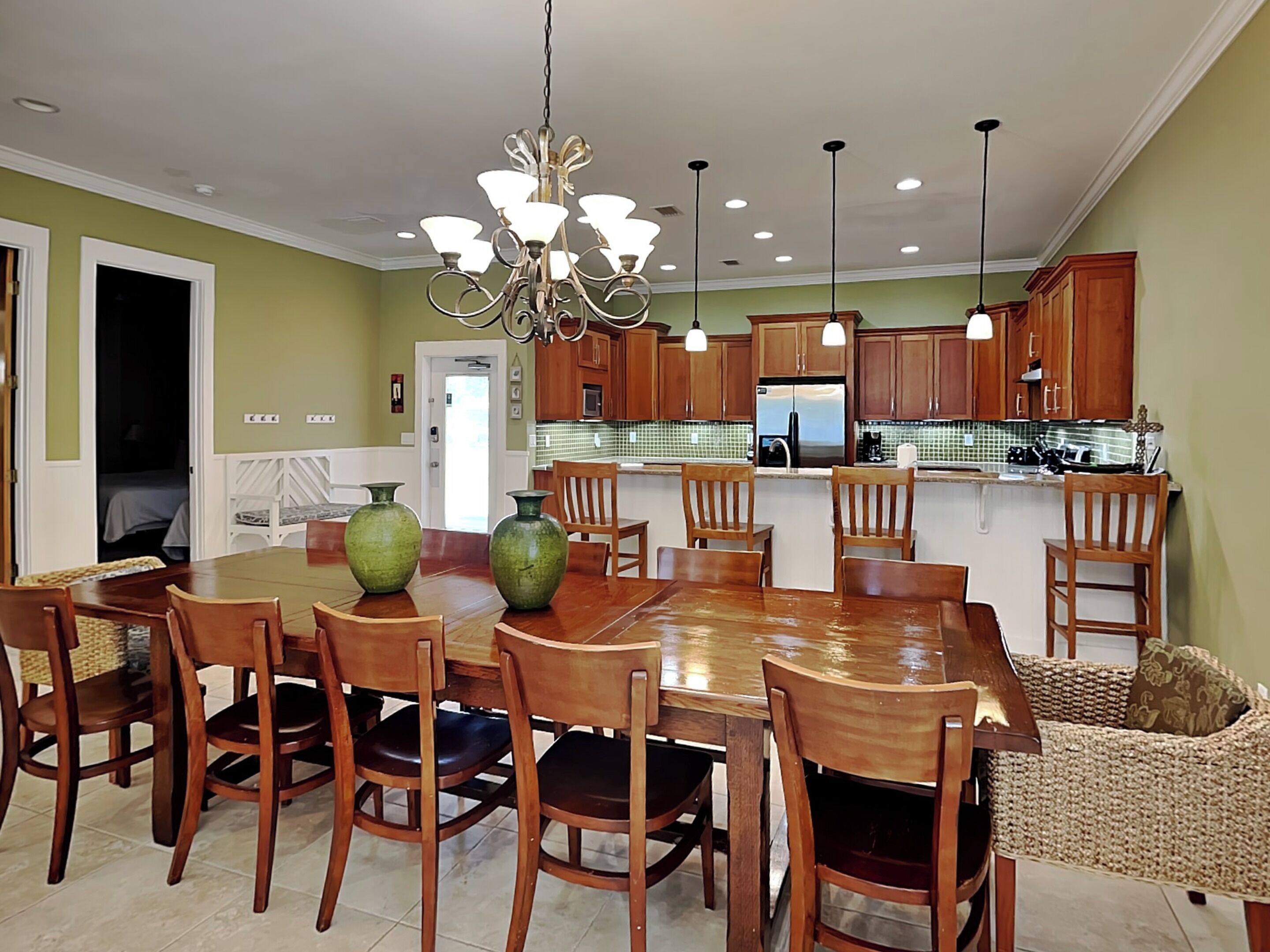 A dining area with a wooden table and chairs, a chandelier, and a kitchen in the background.