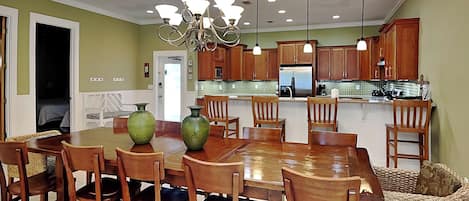 A dining area with a wooden table and chairs, a chandelier, and a kitchen in the background.