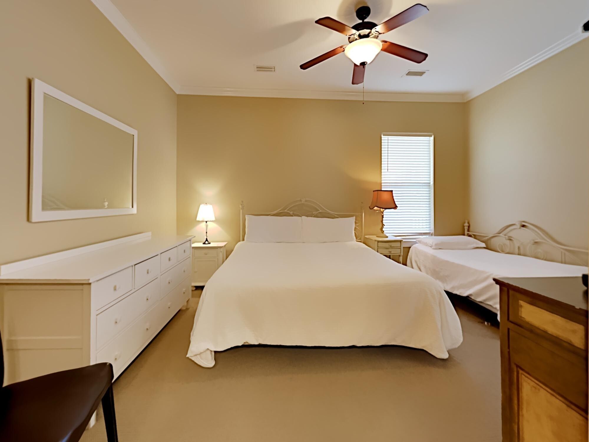 Une chambre à coucher avec un grand lit, deux tables de chevet, un ventilateur de plafond, un miroir et une fenêtre avec des stores.