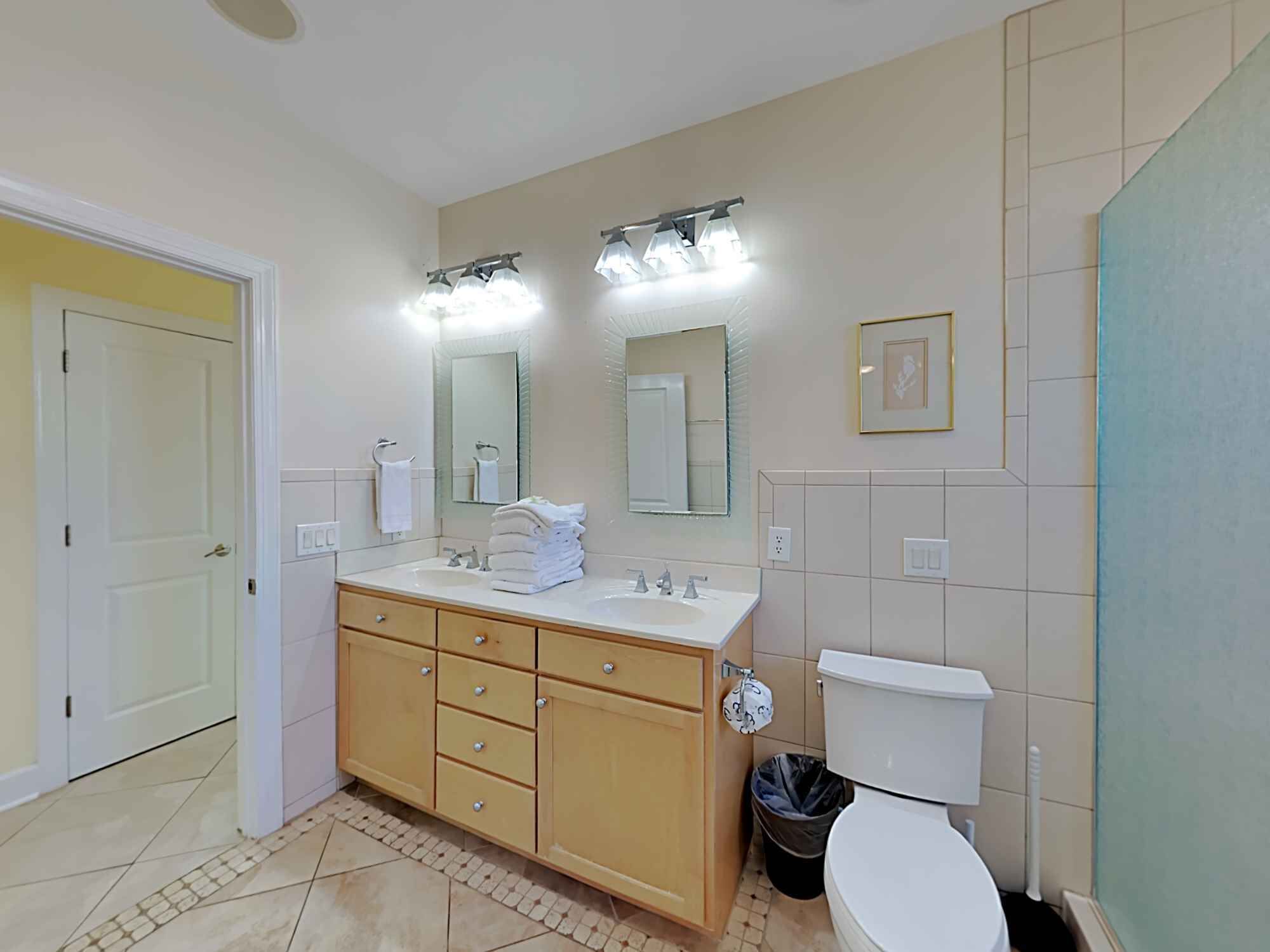 A bathroom with a double sink vanity, a toilet, and a trash bin.
