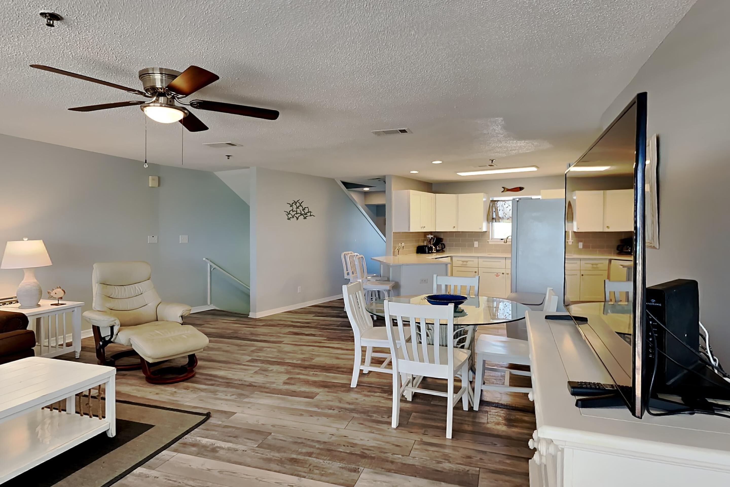 A spacious living room with a ceiling fan, a dining area with white chairs, and a kitchen with modern appliances.