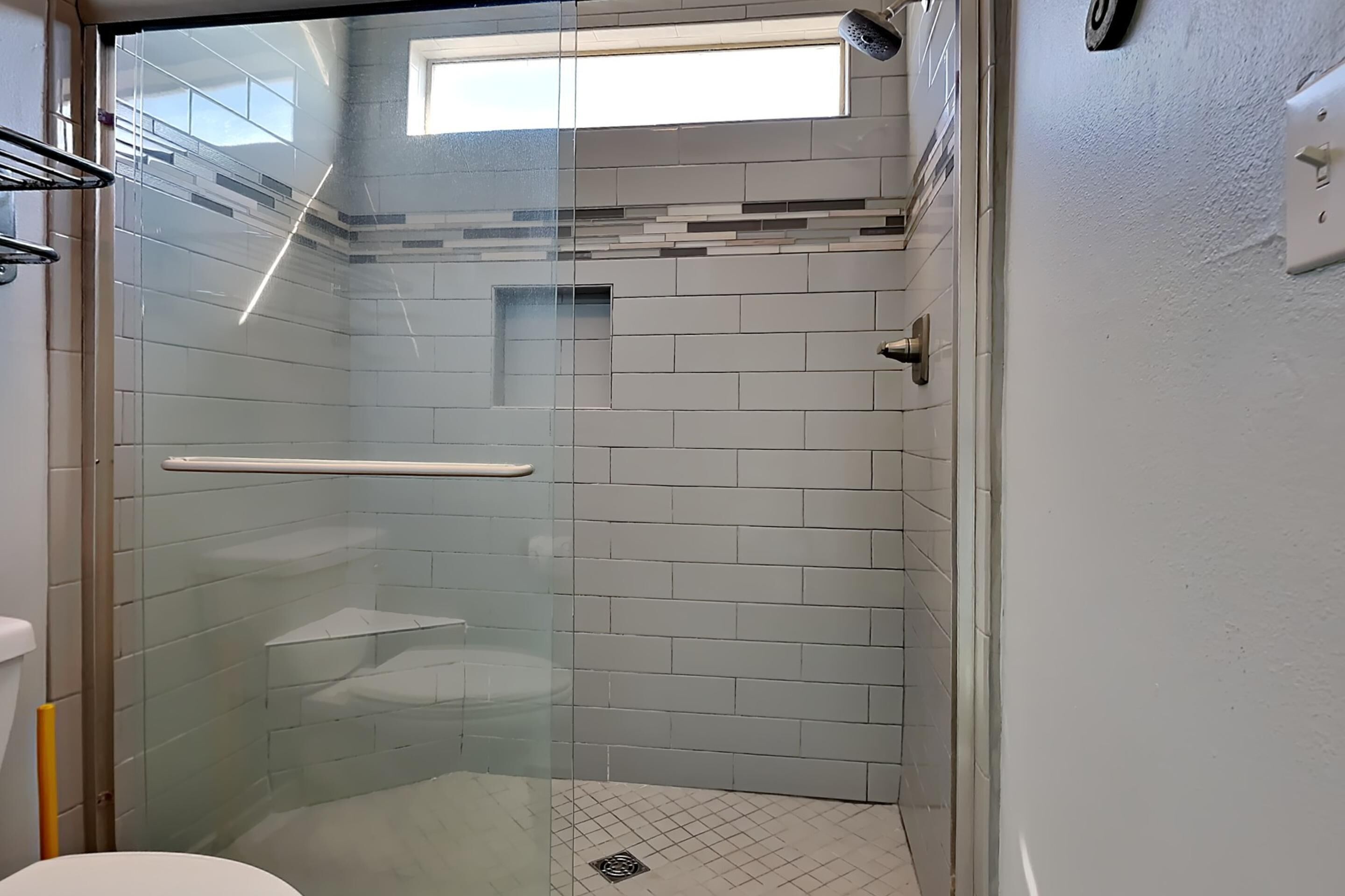 A glass-enclosed shower with a white tiled wall, a glass door, and a small window above the shower.