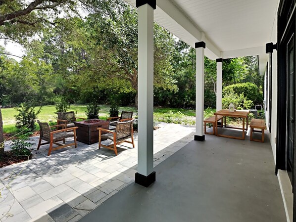 Een overdekt terras met buitenmeubilair, een vuurkuil en uitzicht op een weelderige tuin.