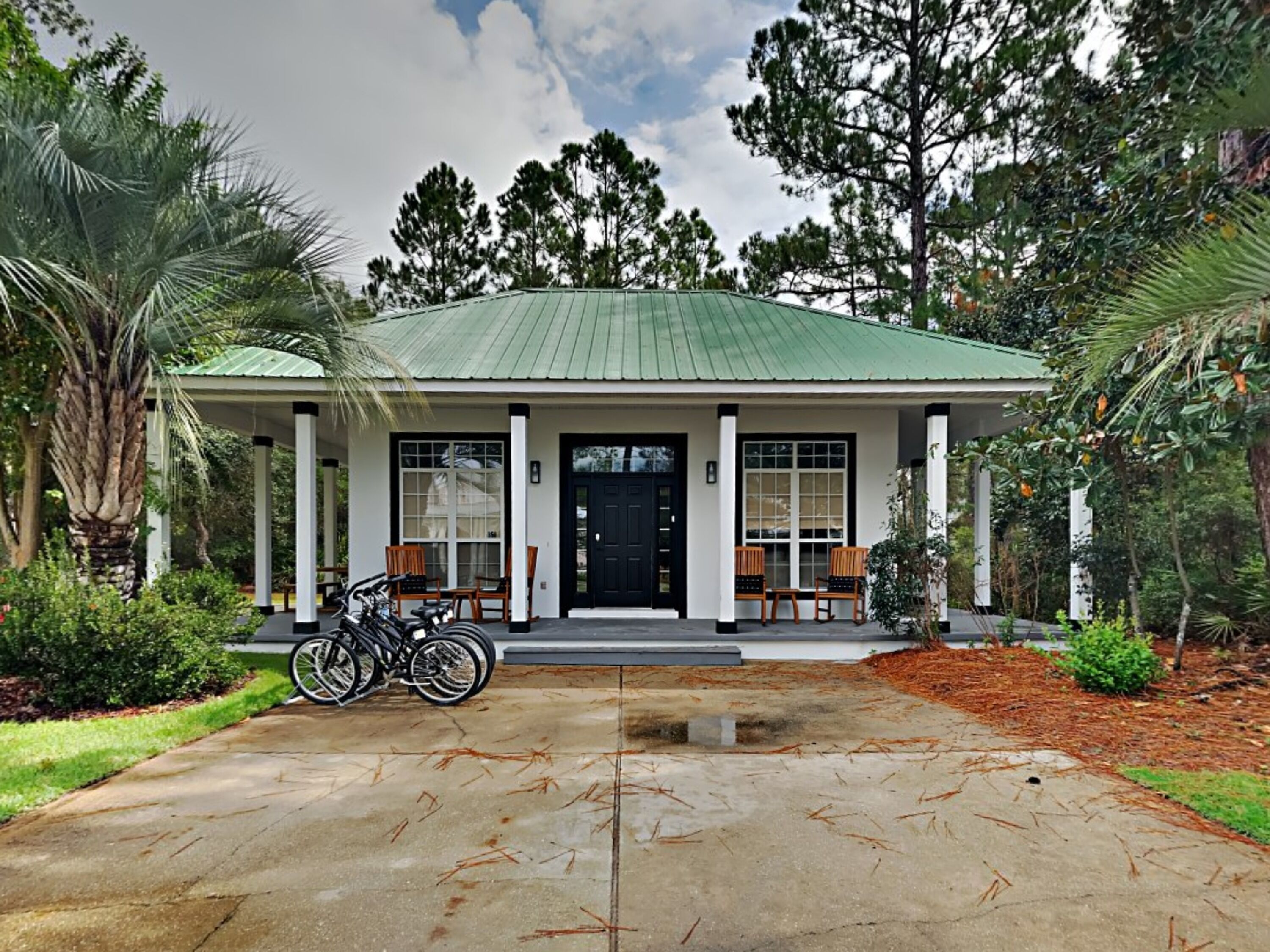 A two-story house with a green roof, white columns, and a black front door. There are two bicycles parked in front, and a covered porch with chairs.