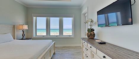A bedroom with a large bed, a dresser, a TV mounted on the wall, and a window with a beach view.