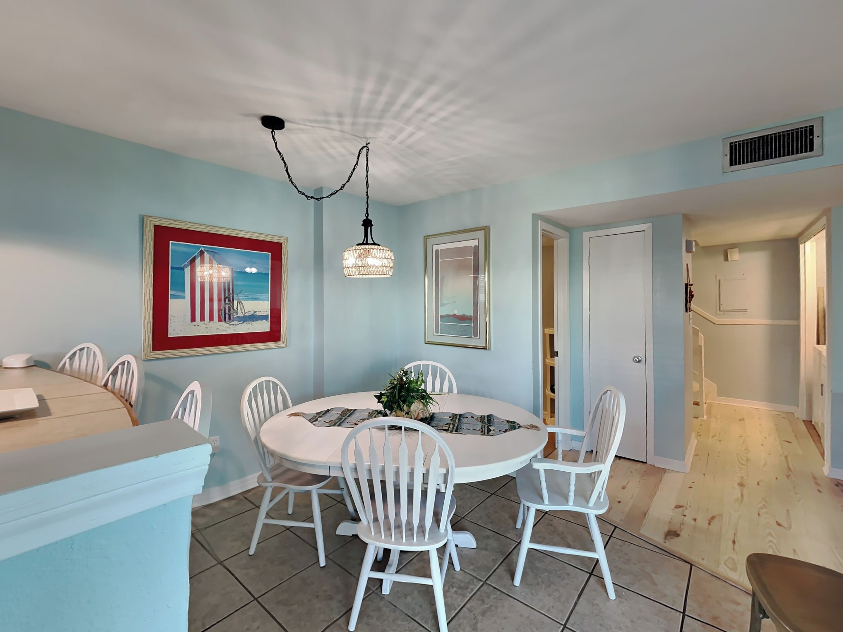 Un coin repas avec une table ronde, des chaises blanches et un ventilateur de plafond. Les murs sont ornés de tableaux encadrés et une porte donne accès à une autre pièce.