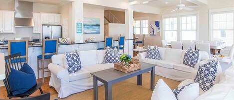 A spacious living area with a white sofa set, blue chairs, and a coffee table. There is a kitchen area with white cabinets and a refrigerator.