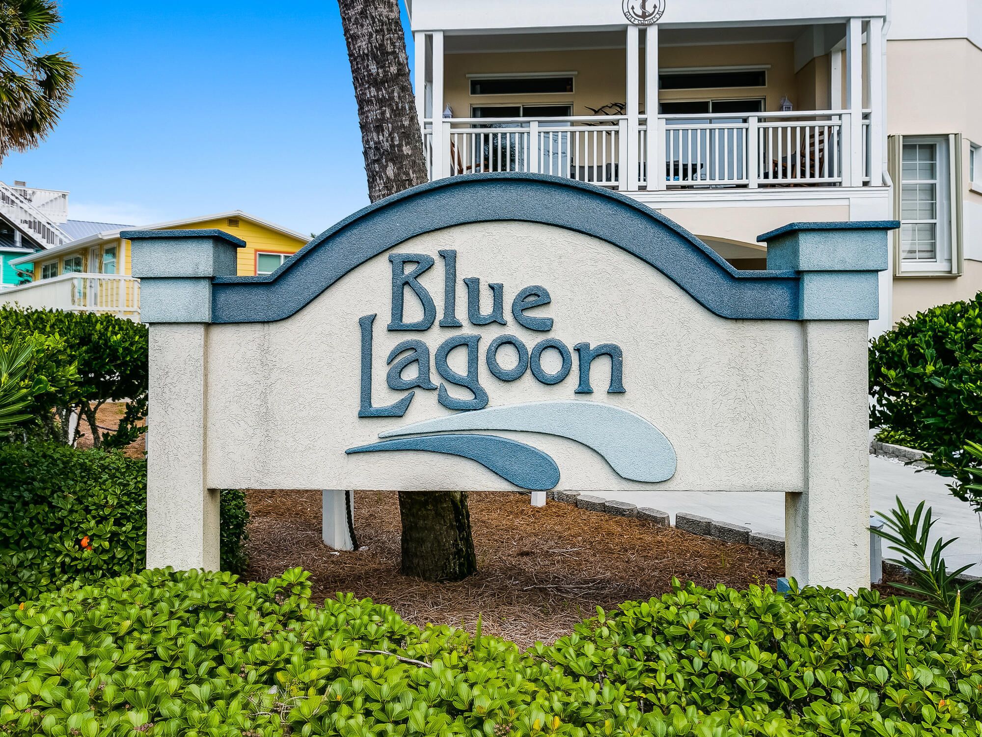A sign for Blue Lagoon with a wave design, surrounded by greenery and a building in the background.