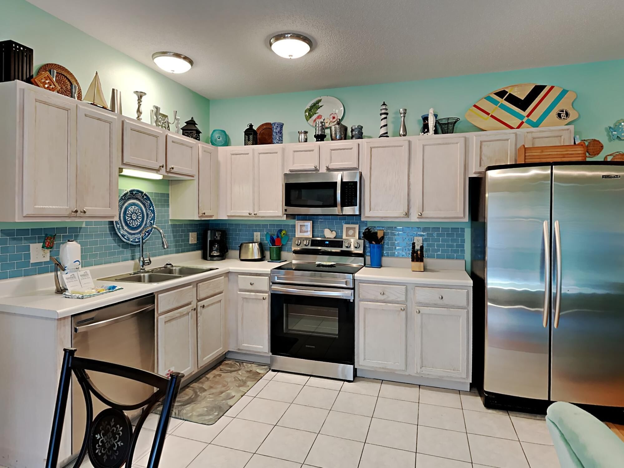 Una cocina con armarios blancos, un refrigerador de acero inoxidable, un microondas y un horno. Hay objetos decorativos en los estantes y un salpicadero azul.