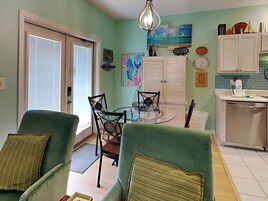 Una habitación con dos sillones verdes, una mesa de comedor con sillas, una zona de cocina con fregadero y armarios, y obras de arte en las paredes.