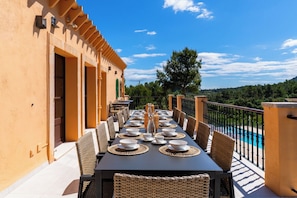 An outdoor dining area with a table set for six, surrounded by wicker chairs, overlooking a swimming pool and a scenic landscape.