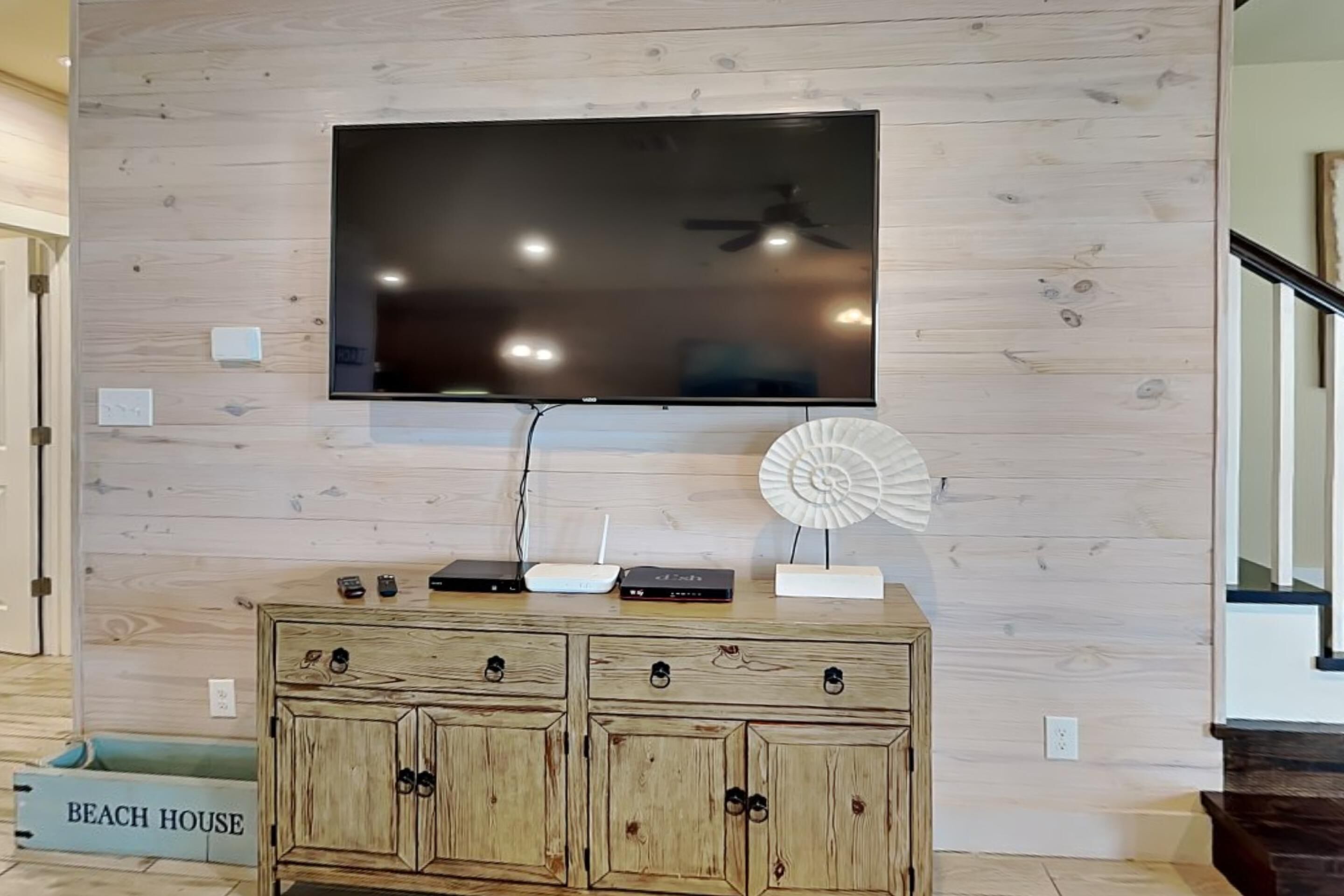 Ein modernes Wohnzimmer mit einem großen Flachbildfernseher an einer Holzwand, einem Holzkasten mit Schubladen und einem kleinen dekorativen Ventilator.