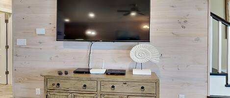 Ein modernes Wohnzimmer mit einem großen Flachbildfernseher an einer Holzwand, einem Holzkasten mit Schubladen und einem kleinen dekorativen Ventilator.