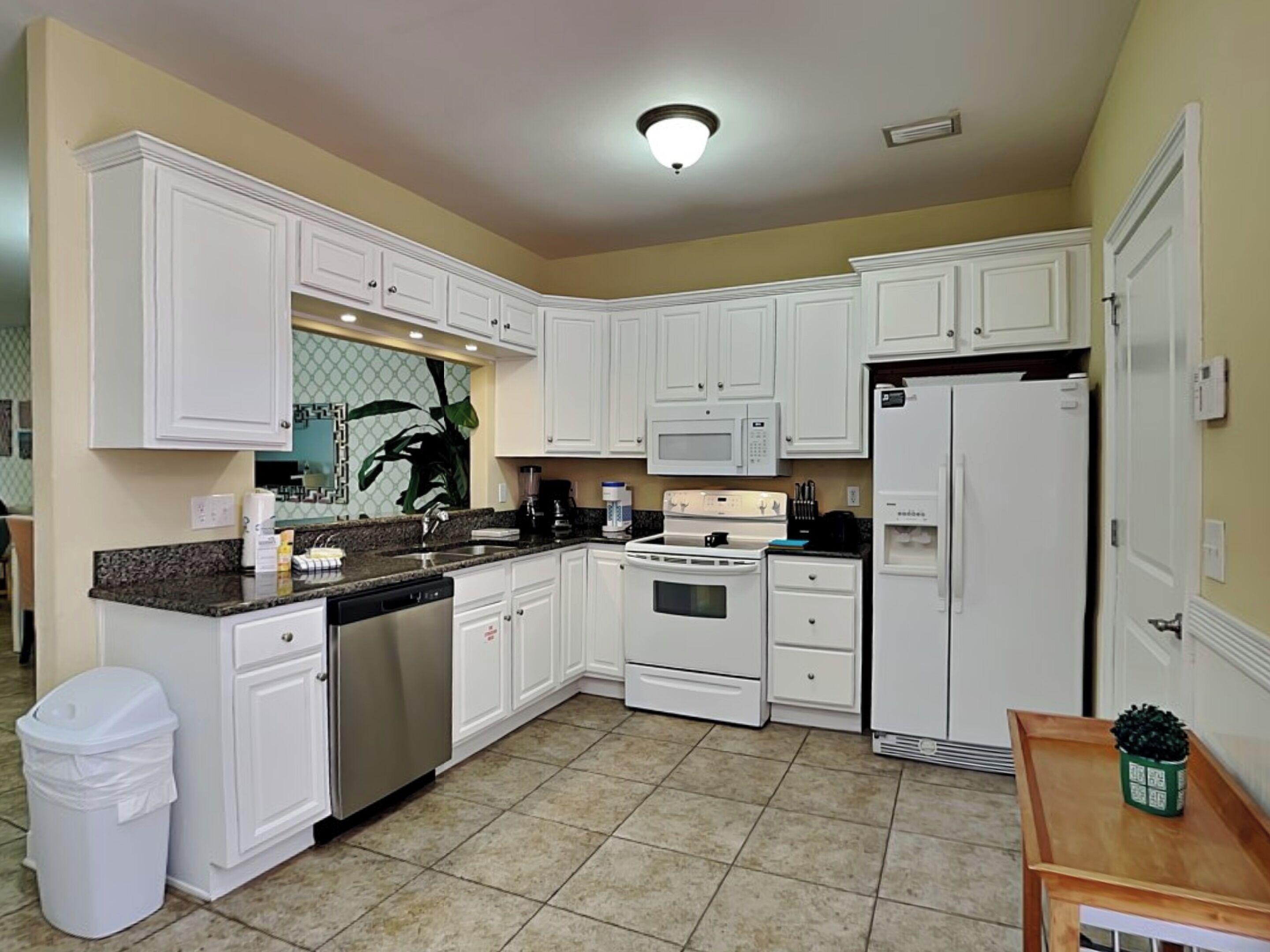 A kitchen with white cabinets, a stove, microwave, and refrigerator. There is a trash can and a small table with a plant.