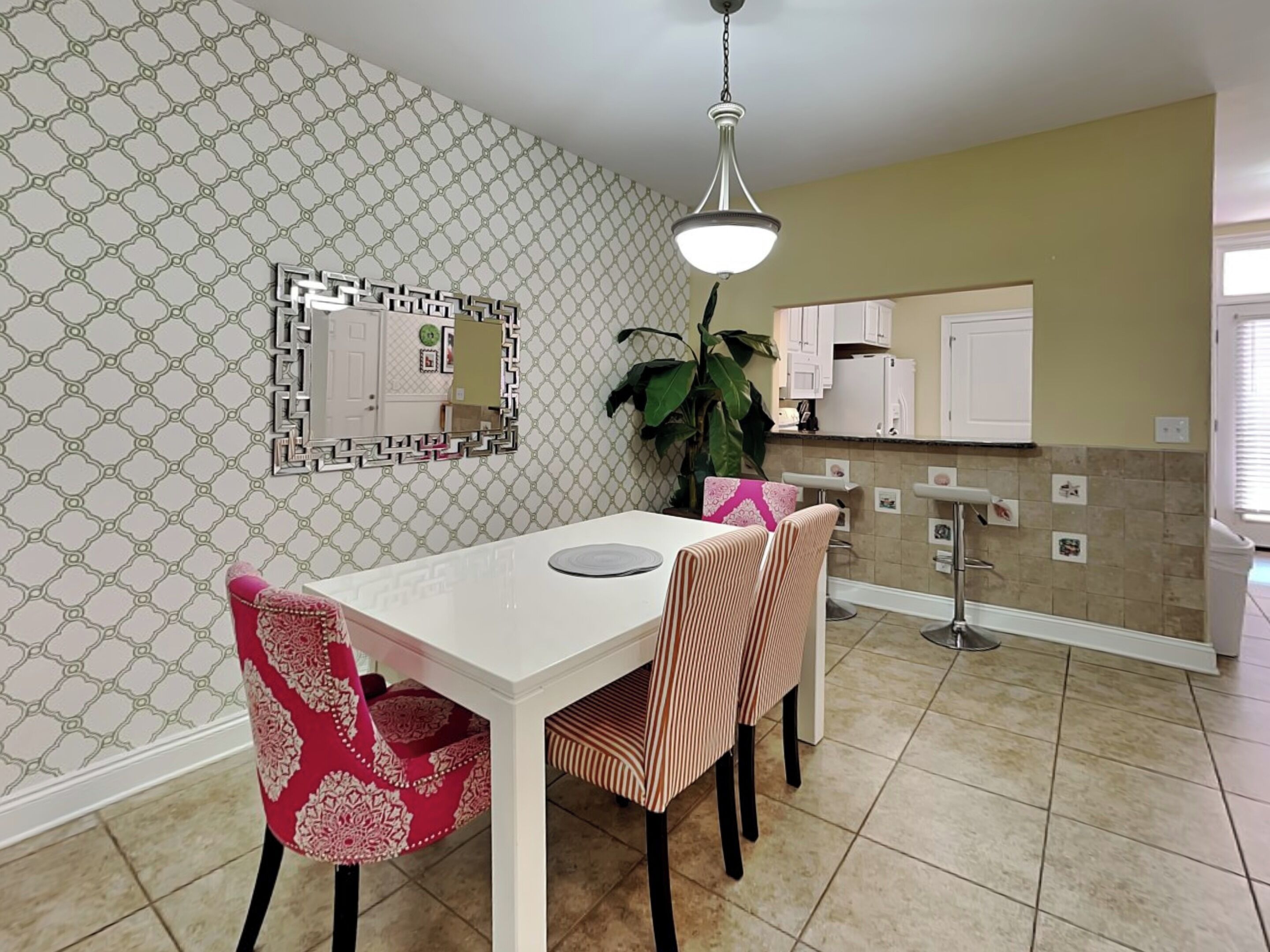 A dining area with a white table and patterned chairs, a hanging light fixture, and a kitchen in the background.
