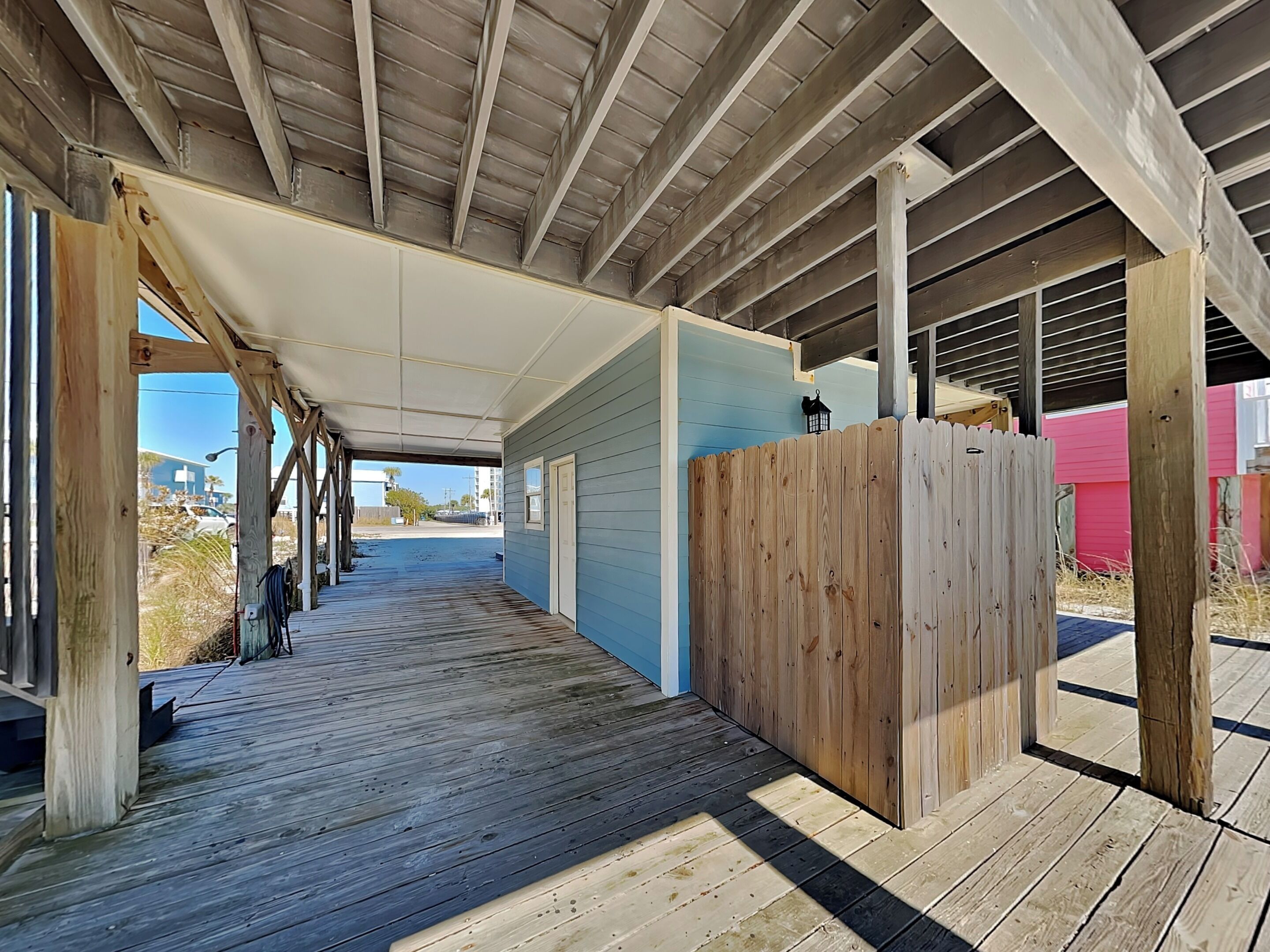 A covered wooden deck with a view of the beach, a wooden fence, and a red building in the background.