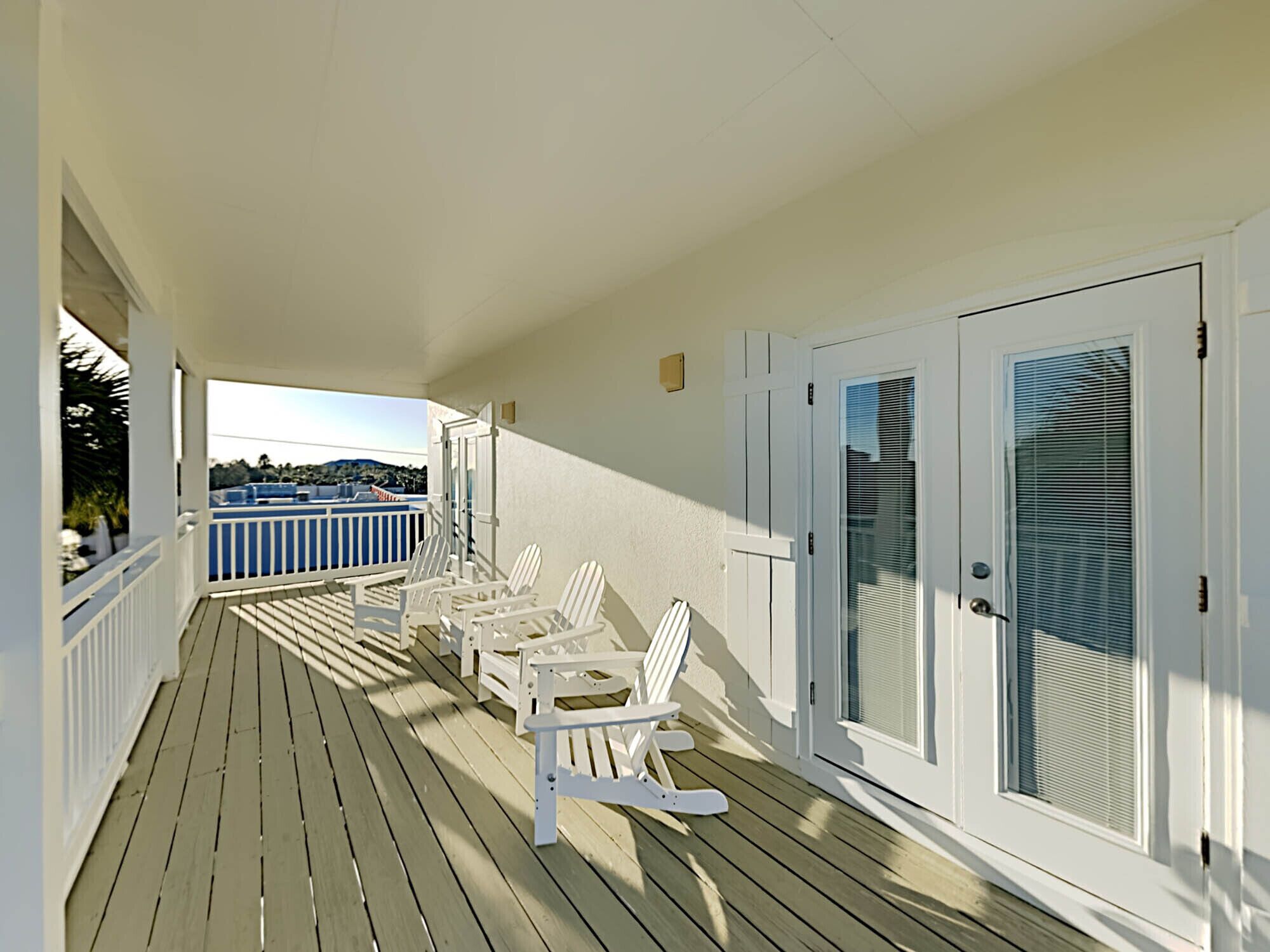 A wooden deck with white chairs and a table, a white door with a window, and a view of a blue railing and distant landscape.