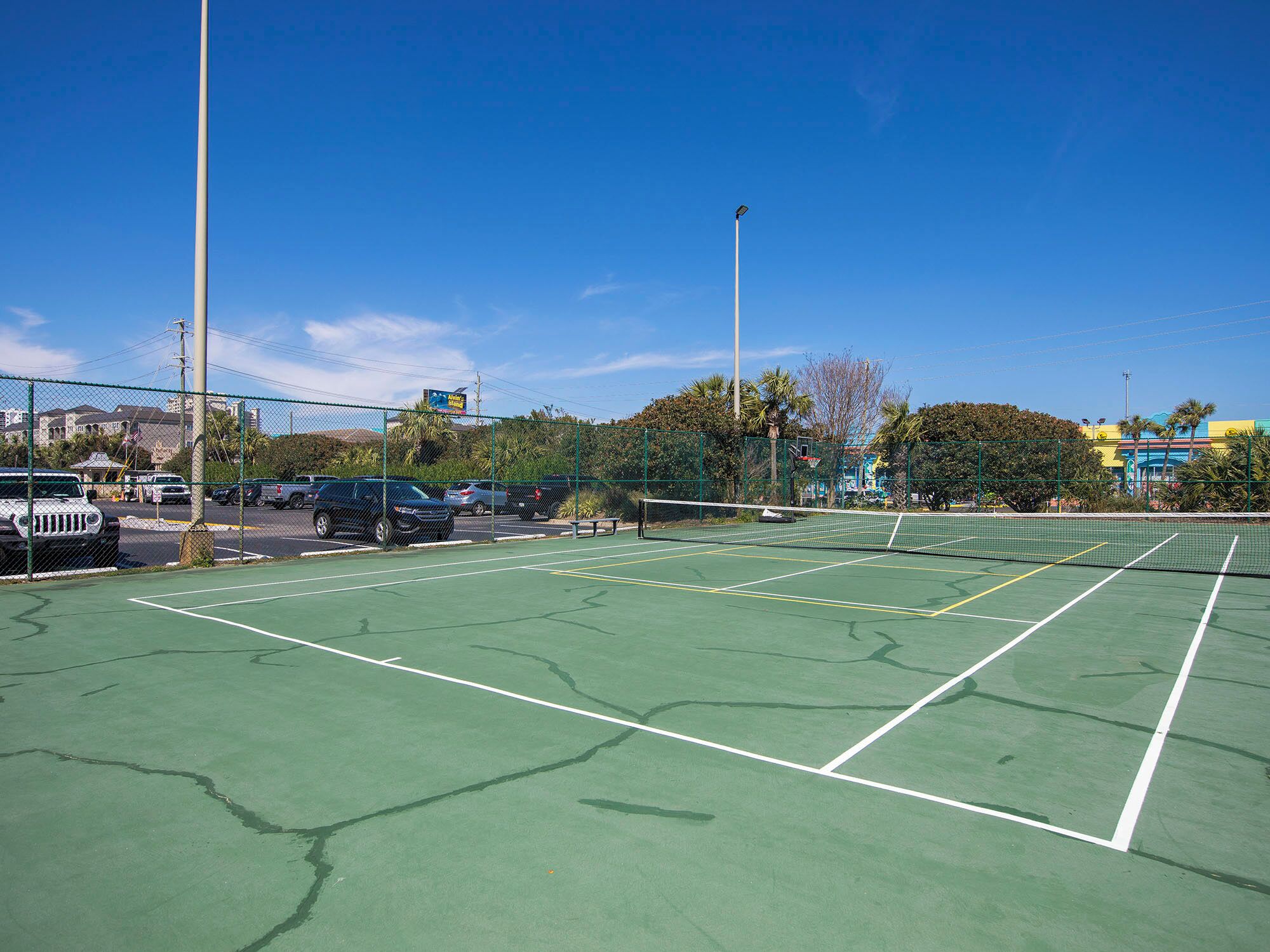 Un court de tennis extérieur, doté d’un filet et entouré d’une clôture, jouxtant un parking où sont garées des voitures.