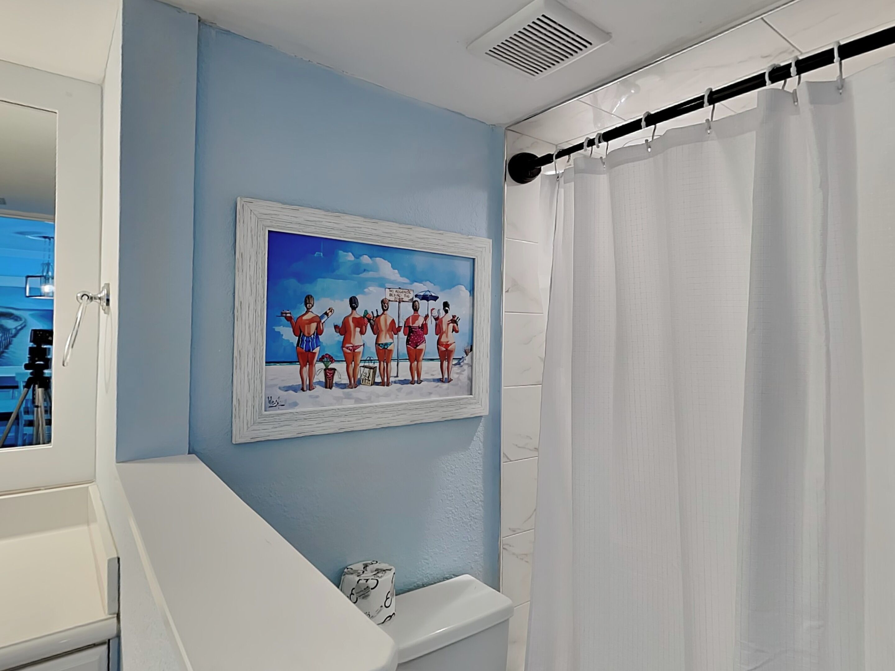 A bathroom with a blue wall, a framed beach scene painting, a white toilet, and a shower curtain.