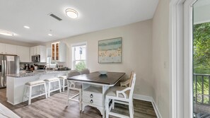 A modern kitchen with white cabinetry, a central island, and a dining area with a wooden table and chairs. - Salt & Sand - Serenity on 30A - Hidden Lakes Condo 321 (Santa Rosa Beach)
