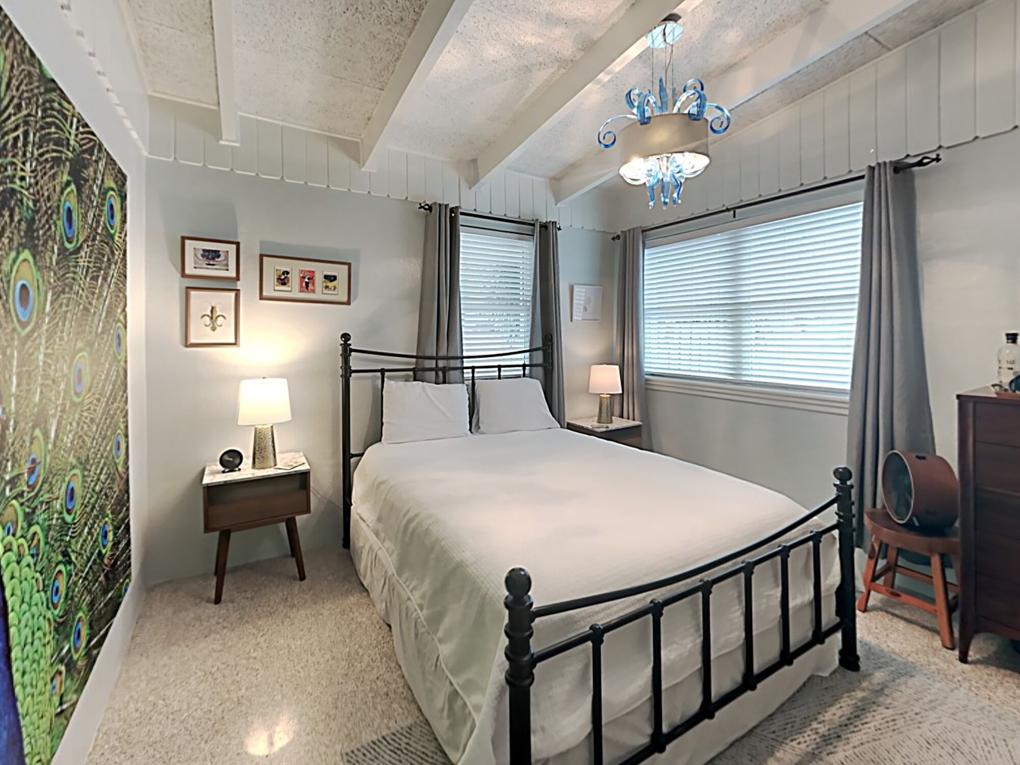 A bedroom with a large bed, two bedside tables with lamps, a chandelier, a peacock-themed wall, and a window with blinds.