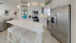 A modern kitchen with white cabinetry, a stainless steel refrigerator, a microwave, and a built-in oven. There is a dining area with a white table and bench. - Aloha Sandy Bay by Southern Vacation Rentals (Santa Rosa Beach)