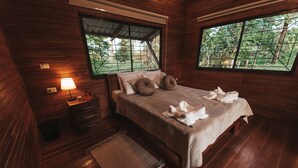 Un dormitorio de cabaña de madera con cama, mesita de noche, lámpara y ventanas amplias con vistas a un bosque.